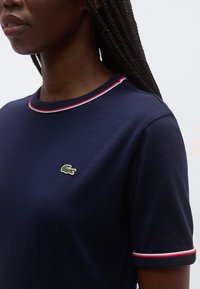 Tumši zila kokvilnas T-krekls ar apaļu kaklu, uz kura redzams neliels zaļš krokodila logo un sarkano un balto apdares lentes apkaklītei un piedurknēm.