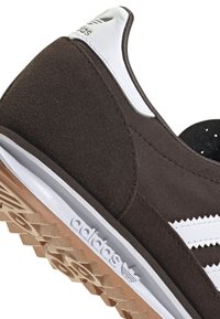 Brown athletic shoe with a suede and fabric upper, white stripes, rubber sole, and embossed logo. Features a textured outsole for grip.