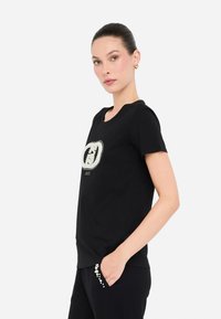 Camiseta negra de manga corta con un logo bordado ornamentado en plata y blanco. Corte ajustado con un cuello redondo y una textura de tela suave.