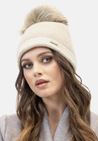 Bonnet beige en tricot avec un large bandeau, orné d'un pompon en fausse fourrure beige sur le dessus et d'un petit logo en argent sur le côté.