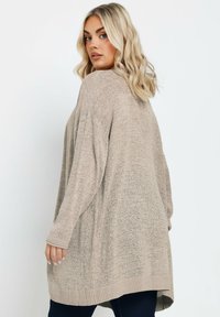 Beige, löst sittande tröja med en texturerad stickad design, långa ärmar och ribbad fåll, visad bakifrån med en rundad form.