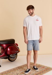 T-shirt en coton blanc avec un graphique rouge imprimé "PIZZA" sur la poitrine, associé à un short en denim bleu clair et des baskets marine. Scooter vintage en arrière-plan.