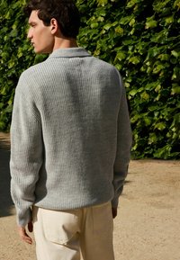 Grauer, gerippter Strickpullover mit Kragen, lockere Passform, lange Ärmel. Kombiniert mit hellbeigen Hosen, vor einem grünen, blätterreichen Hintergrund.