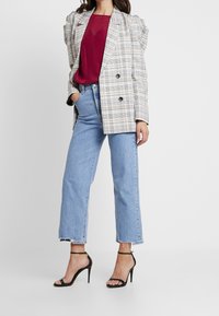 Blazer doppiopetto in plaid grigio con spalle strutturate, abbinato a un top bordeaux e jeans a vita alta di colore blu chiaro, e sandali neri con tacco alto.