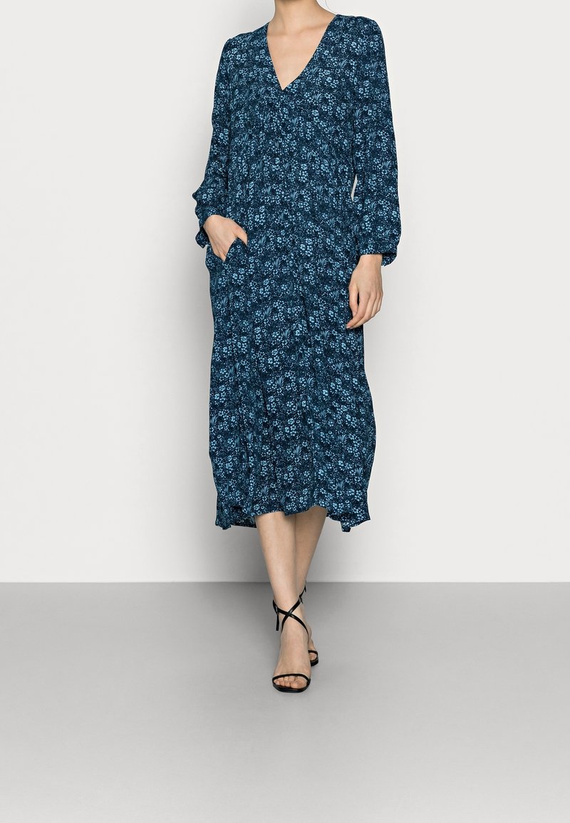 Femme portant une robe midi fleurie bleu marine à manches longues avec col en V et poches, associée à des sandales noires à lanières.