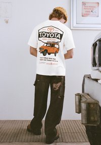 Biała koszulka z nadrukiem Toyoty Land Cruiser w kolorach czarnym i pomarańczowym, zestawiona z czarnymi, postrzępionymi spodniami oraz brązowym obuwiem.