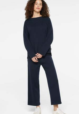 Frau mit lockigem Haar, die ein marineblaues langärmliges Oberteil und weit geschnittene Hosen trägt, steht mit verschränkten Händen, weiße Turnschuhe, weißer Hintergrund.