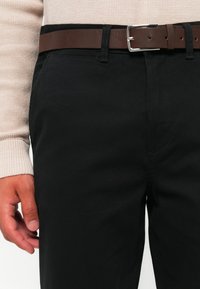 Man wearing black pants with a brown leather belt and a beige textured long-sleeve sweater, hand relaxed by his side.