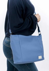 Blaues, strukturiertes Handtasche mit zwei Seitentaschen, verstellbarem Tragegurt und Logo-Tag. Das Material scheint strapazierfähig zu sein, mit rechteckiger Form.