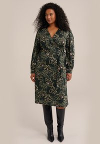 Rochie tip wrap din material verde închis, cu modele florale în tonuri de aur și violet. Mâneci lungi, talie accentuată și tiv până la genunchi. Botine negre până la genunchi.