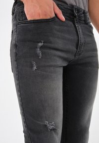 Primer plano de una persona que lleva jeans negros desgastados, con la mano en el bolsillo delantero, contra un fondo liso claro.