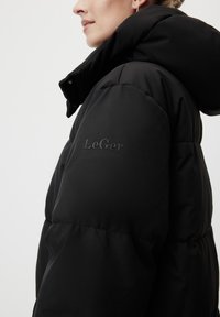 Femme portant une doudoune noire avec capuche, logo de la manche "LeGer" visible, montrée de l'épaule au menton en profil latéral sur fond blanc.