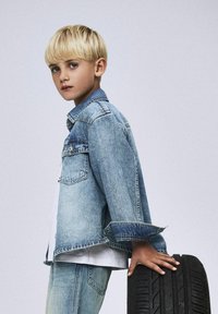 Helehall denim jakk valge särgi ja siniste teksadega, mille varrukad on üles keeratud, toetades kätt mustale rehvi külge.