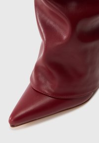 Acercamiento a una bota de tobillo de cuero roja oscura, de punta afilada y caída, sobre fondo blanco.