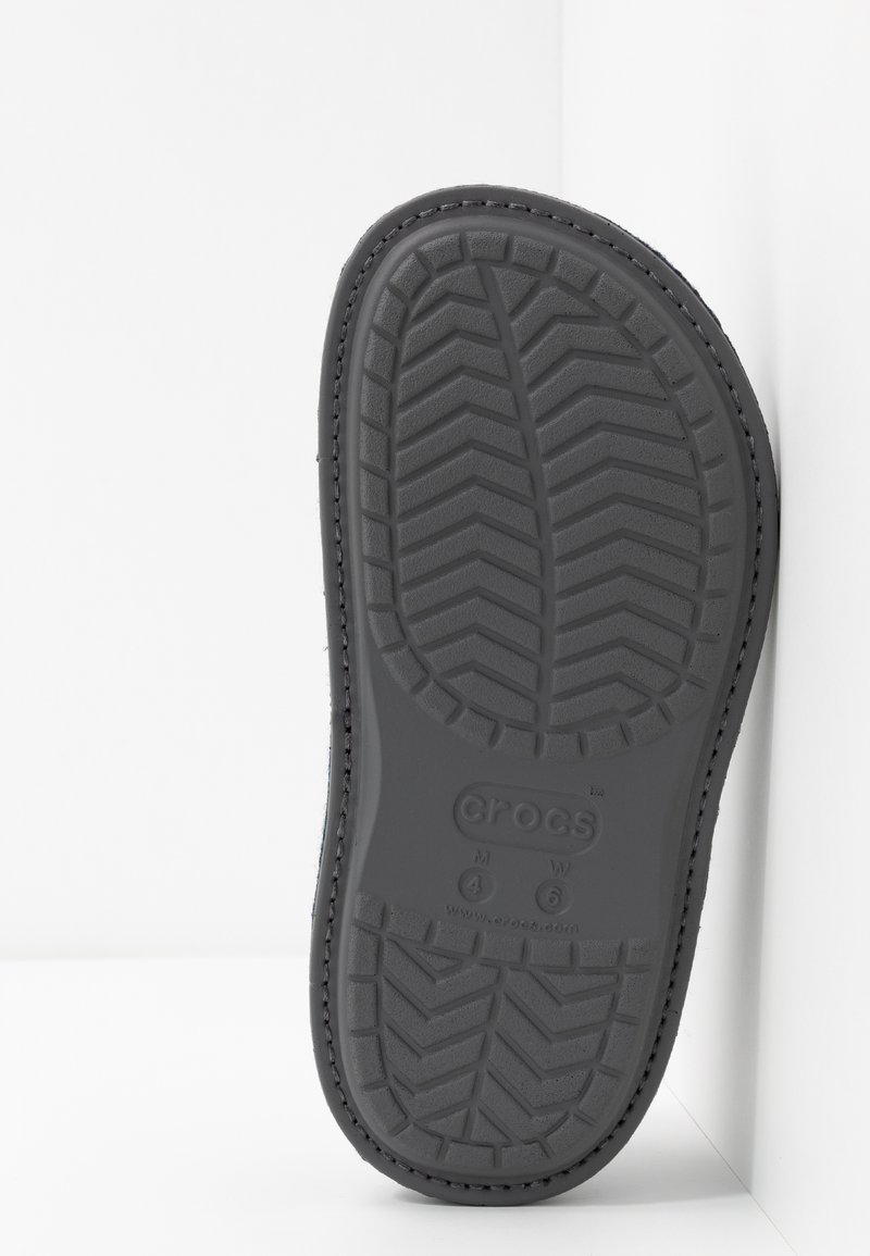 Suela de Crocs de goma negra con un patrón de rodadura texturizado, que presenta agarres elevados y bordes suaves, mostrando el logotipo de la marca y marcas de tamaño.