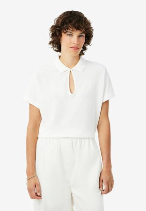 Vrouw met krullend haar die een wit topje met korte mouwen en een kleine sleutelgatovergang aan de voorkant draagt, gecombineerd met witte broek met hoge taille, tegen een effen achtergrond.