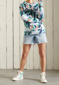 Persona vistiendo una sudadera con estampado floral, pantalones cortos de mezclilla azul claro desgastados y zapatillas blancas de caña alta, de pie contra una pared de paneles de madera.