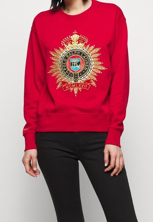 Rød sweatshirt med gyldent broderet Polo Ralph Lauren våbenskjold og romertal, båret med sorte bukser af en stående person.