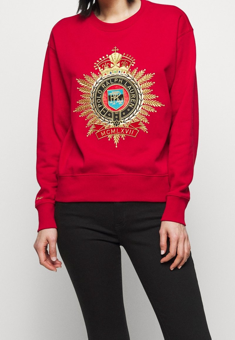 Sweat-shirt rouge avec blason Polo Ralph Lauren brodé en or et chiffres romains, porté avec un pantalon noir par une personne debout.