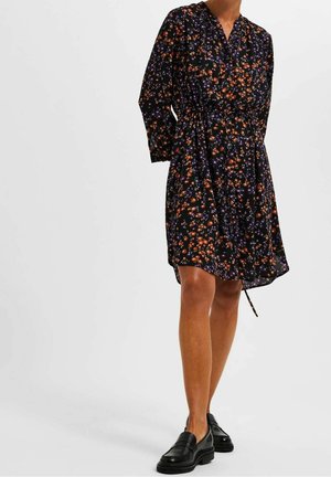 Czarna sukienka kwiatowa z pomarańczowymi i fioletowymi kwiatami, długie rękawy, dekolt w serek i wiązany w talii; warstwowo zestawiona z czarnymi, masywnymi mokasynami.