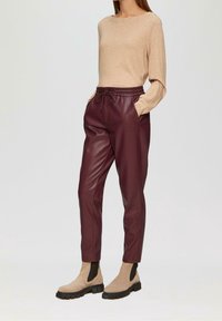 Žena v béžovém dlouhém rukávu, s kalhotami z burgundské umělé kůže s vysokým pasem a šňůrkou, a béžovými kotníkovými botami s černými elastickými bočnicemi.