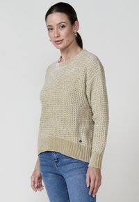 Beež kampsun, millel on tekstuuriga muster, soonikkoes kaelus ja äär, pikad varrukad ning mugav istuvus. Kombineeritud siniste teksadega.