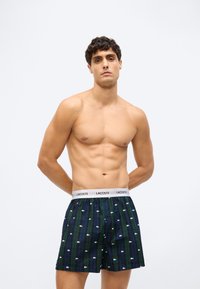 Bărbat fără tricou, cu păr creț, purtând boxeri Lacoste în carouri verzi și bleumarin, cu un tiv alb, pe un fundal simplu deschis.