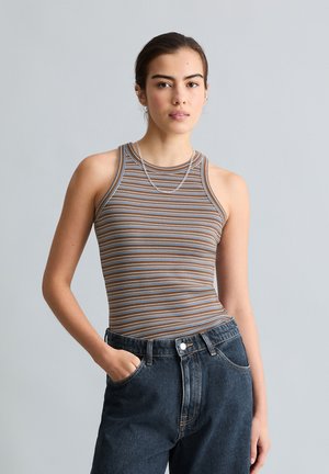 Jeune femme portant un top sans manches rayé marron et bleu et un jean taille haute foncé, debout avec une main dans la poche sur fond gris.