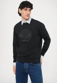 Czarna bluza z teksturowanym logo na środku, w duecie z niebiesko-białą koszulą w paski pod spodem. Całość dopełniają dżinsowe spodnie.