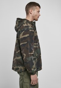 Camouflagetjack in tinten van groen en bruin, met een capuchon, elastische manchetten en een ritszak aan de zijkant. Lichtgewicht materiaal.
