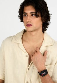 Orologio smartwatch nero con una faccia rettangolare, accenti rossi e bianchi sul display, indossato al polso di una persona con una camicia button-up color crema.