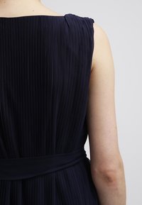 Sinivihreä pleatattu mekko, jossa on olkaimeton muotoilu, vyötärönauha ja sileä rakenne. Kangas on kevyttä ja siinä on pystysuorat poimut.