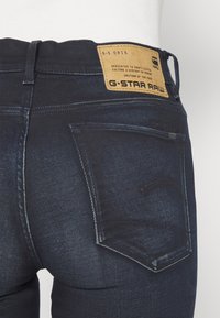 Jeans de mezclilla azul oscuro con corte slim, que presentan una costura elevada en el bolsillo trasero, una etiqueta de cuero en la cinturilla y una sutil textura desgastada.