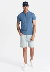 Modrá polo košile s logem, světle modré denimové šortky a bílé tenisky. Model má na sobě námořnickou čepici s bílým nápisem.