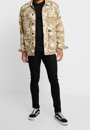Man wearing beige and white camo jacket over black shirt, black skinny jeans, and black and white sneakers standing on white background.