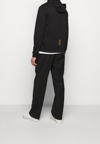 Svart huvtröja med en texturerad fåll, gyllene vertikala ränder på baksidan, ihopparad med lösa svarta byxor och vita sneakers.