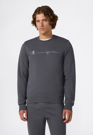 Joven vestido con un sudadera gris oscura de Champion y pantalones deportivos a juego, de pie contra un fondo blanco liso.
