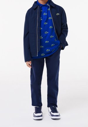 Veste zippée navy sur un polo bleu avec des motifs de crocodile verts, associée à un pantalon navy et des baskets blanches/navy.