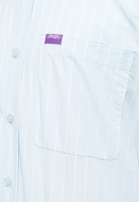 Gai zils svītrots krekls, izgatavots no kokvilnas, ar kabatu un purpura "JACKER" etiķeti uz krūts. Iezīmes: pogu aizdare priekšpusē un maiga tekstūra.