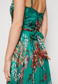 Floral dress in teal fabric, featuring a fitted bodice, spaghetti straps, and a bow accent at the waist. Vibrant floral patterns in reds and whites.