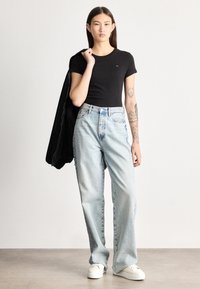 Ung kvinde med tatoveringer iført sort t-shirt, lyseblå viddebukser, hvide sneakers, med sort jakke over skulderen, stående på grå gulv.