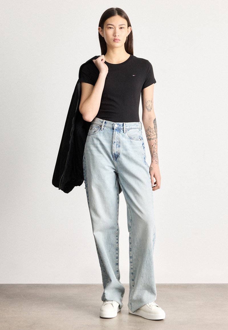 Ung kvinde med tatoveringer iført sort t-shirt, lyseblå viddebukser, hvide sneakers, med sort jakke over skulderen, stående på grå gulv.