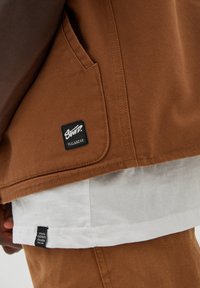 Braune Leinenjacke mit einem sichtbaren schwarzen Markenpatch, die über einem weißen Hemd getragen wird. Verfügt über eine versteckte Tasche und gestickte Akzente.