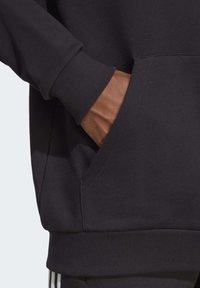 Felpa nera con tasca a marsupio, polsini a costine e una texture liscia. Ingrandimento che mostra la posizione della mano nella tasca.