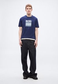 Mornarsko modra bombažna majica s prelivnim logotipom »BOSS«, združena z ohlapnimi temnimi kavbojkami in črnimi čevlji. Enostavna in sodobna zasnova.