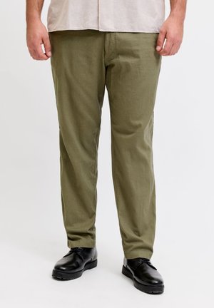 Man wearing olive green pants and black lace-up shoes standing against a plain white background.