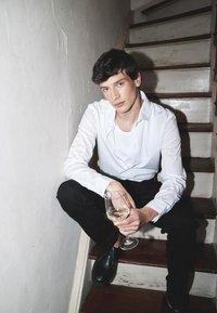 Balta pogu krekls, melni bikses, melni apavi; sēž uz kāpnēm, tur rokās vīna glāzi; gludās tekstūras un pieslāpētas krāsas.