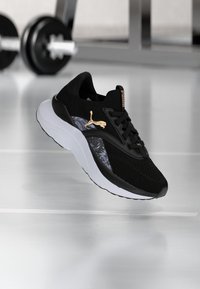 Schwarzer Sportschuh mit Mesh-Obermaterial, goldenem Logo, weißer Sohle und gemustertem Seitenpanel. Verfügt über ein strukturiertes Profil und ein Schnürsystem.