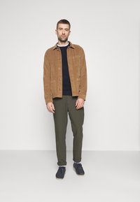 Jachetă de catifea maro cu buzunar frontal, pulover bleumarin dedesubt, pantaloni verde măsliniu și pantofi bleumarin cu o talpă deschisă. Modelul stă pe un fundal simplu.