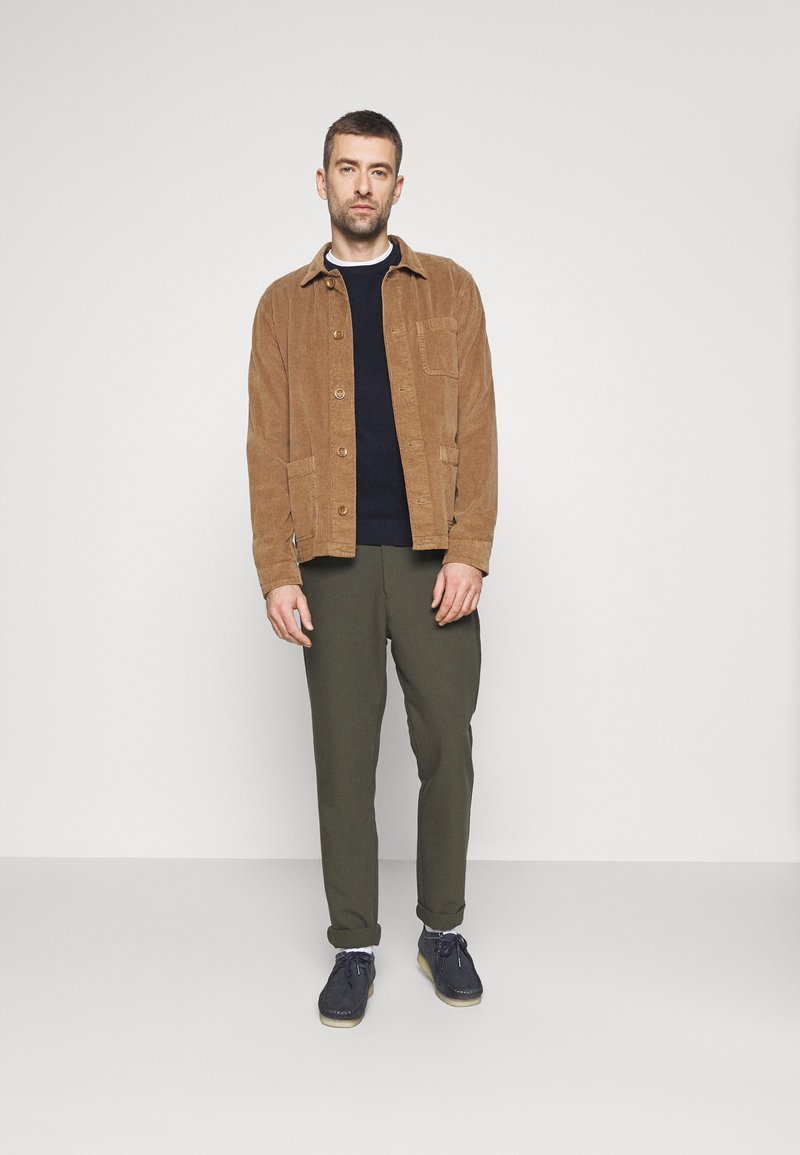 Chaqueta de pana marrón con bolsillo frontal, suéter azul marino debajo, pantalones verde olivo y zapatos azul marino con suela clara. El modelo está de pie contra un fondo sencillo.